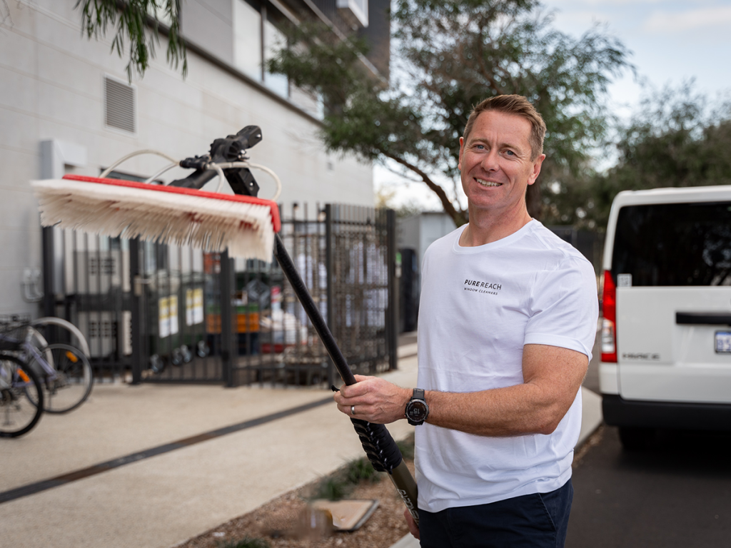 Richard, the owner of Pure Reach Window Cleaning, holding a water-fed cleaning brush, standing outdoors near a commercial property.